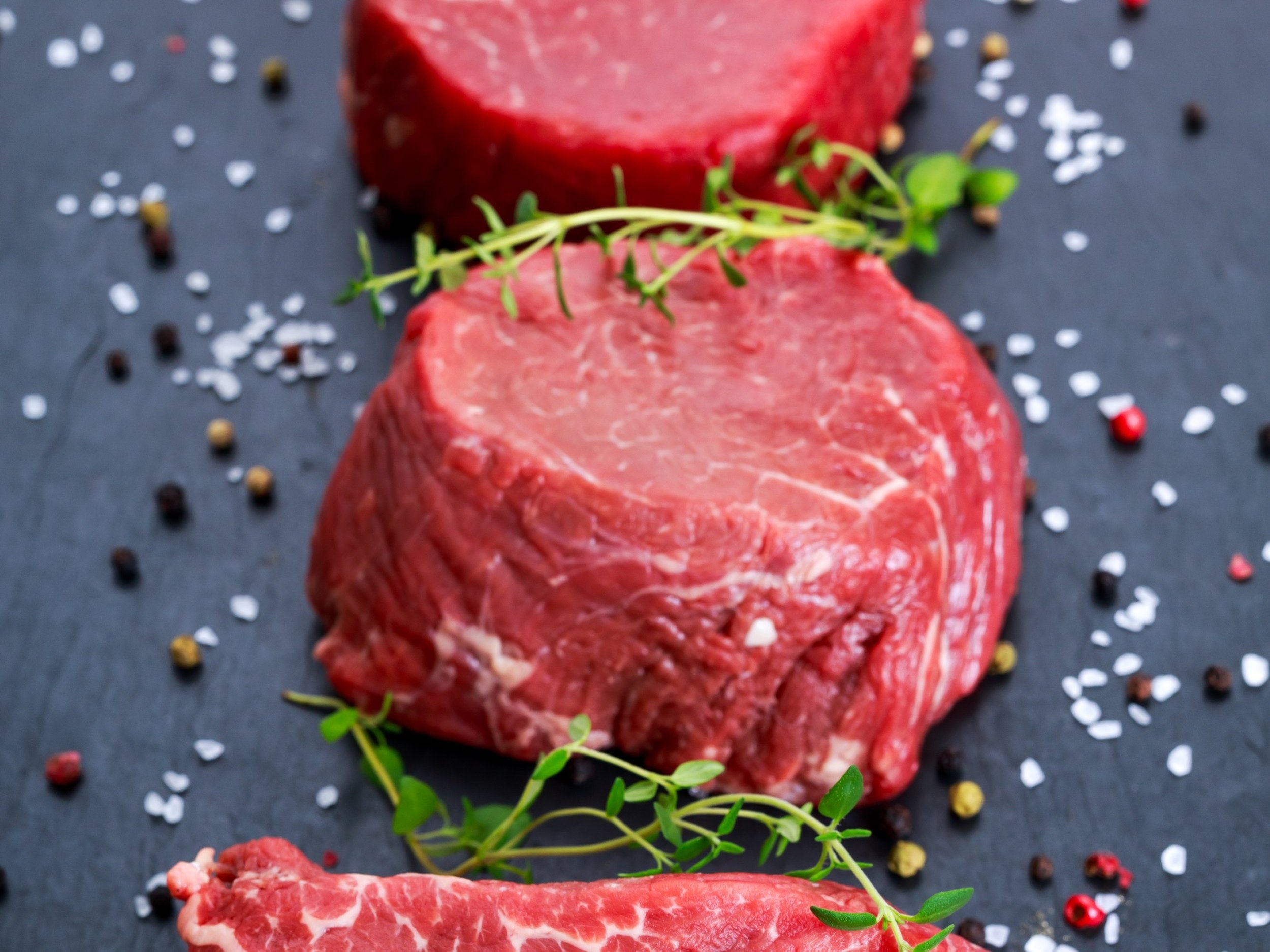 beef_filet Assorted cuts of fresh beef steak garnished with herbs on a dark slate background, surrounded by colorful peppercorns and sea salt.