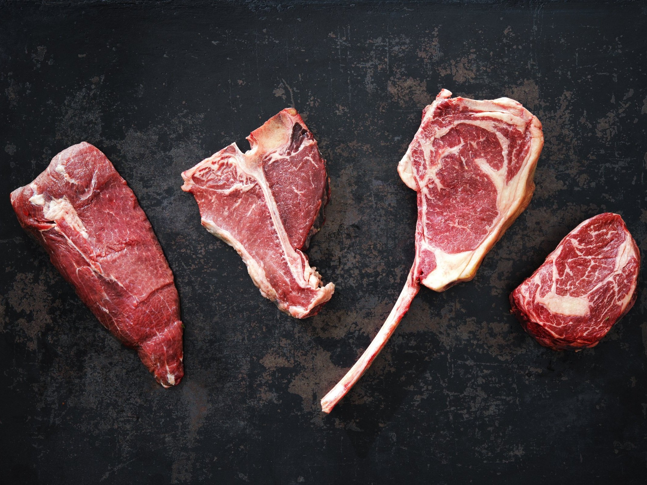 beef_mixed_porterhouse_ribeye_tenderloin Assorted cuts of raw beef including ribeye, T-bone, and filet mignon displayed on a dark textured surface, highlighting the marbling and freshness of the meat.